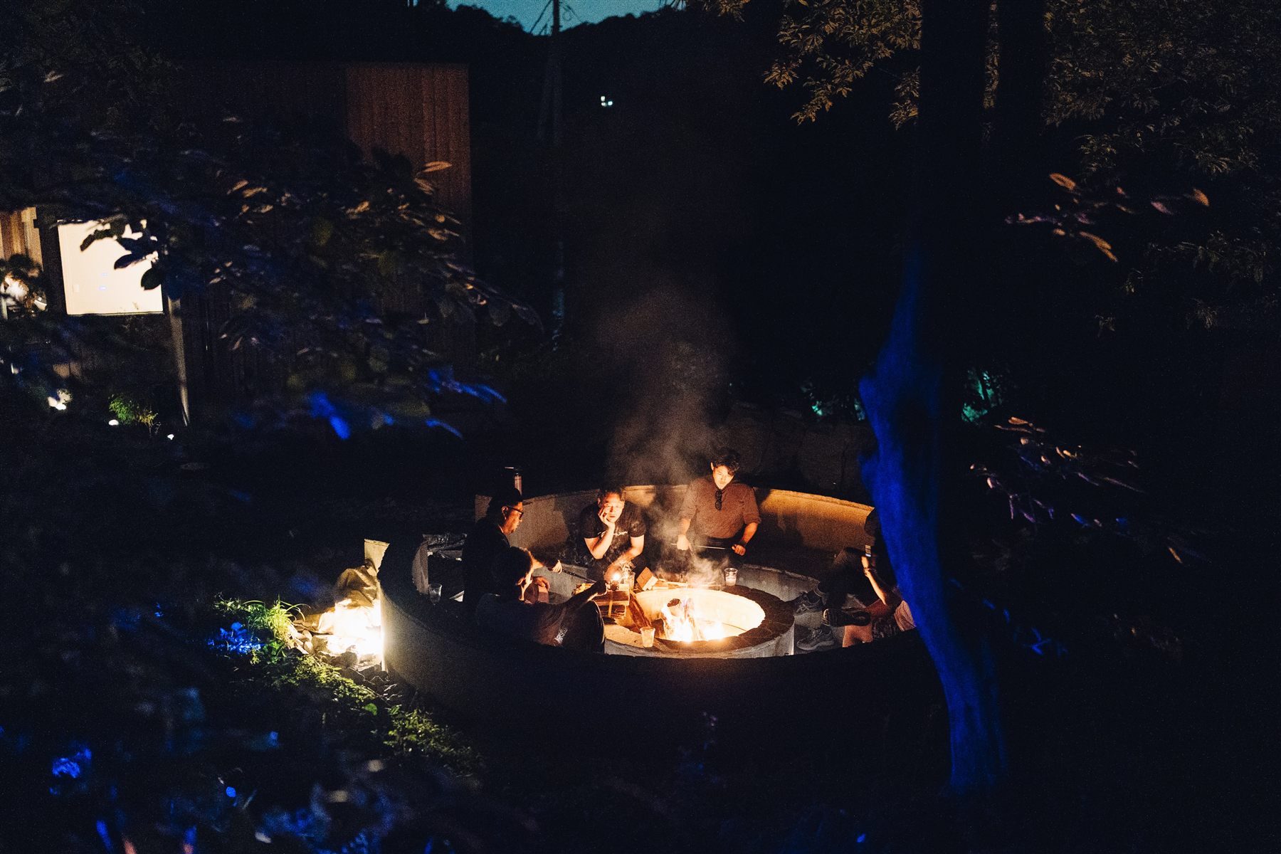 夜の森の中、木々の向こうに焚火と煙が見える